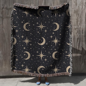 May include: A black woven throw blanket with a repeating pattern of gold crescent moons and stars. The blanket has fringe on all sides.