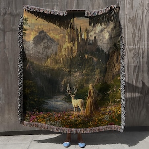 May include: Woven tapestry blanket featuring a fantasy landscape with a castle, deer, and a woman in a golden dress. The blanket has a fringed edge and depicts a scenic view of a river, mountains, and colorful flowers.