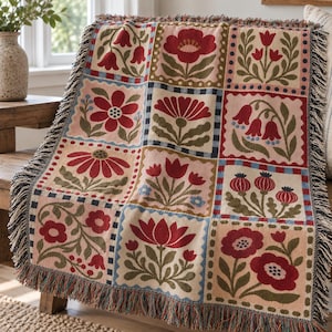 May include: A woven throw blanket featuring a grid of square panels, each displaying distinct floral illustrations in shades of red, olive green, and cream. The blanket is finished with a decorative fringe along the edges and a checkered border.