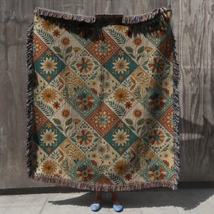 Op de afbeelding: Een geweven sprei met een bloemenpatroon in tinten teal, oranje en beige. Het ontwerp heeft een herhalend vierkant patroon met bloemen en bladeren. De deken heeft een gefranjerde rand.