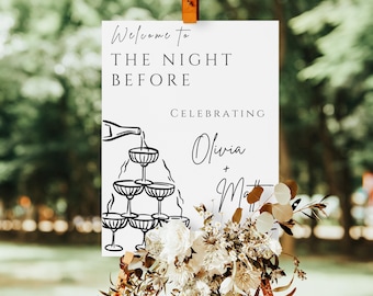 May include: A white sign with the text "Welcome to THE NIGHT BEFORE Celebrating Olivia + Matt" and a champagne tower illustration. The sign is decorated with white and brown flowers and foliage.