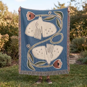 May include: A rectangular woven throw blanket in a deep blue color featuring two large cream-colored birds with spread wings. The design includes long green stems, dark green leaves, and two pinkish-red flowers. The edges have a fringed trim.