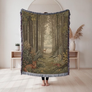May include: A woven tapestry blanket depicting a forest scene with a path, deer, and foxes. The blanket has a fringed edge with green, brown, and beige tones. The artwork shows a tranquil woodland landscape.