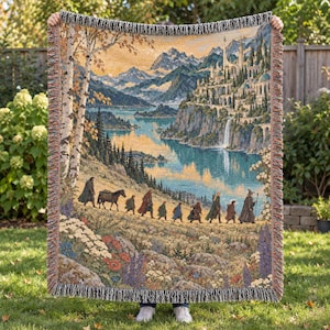 May include: A woven throw blanket featuring a detailed tapestry scene of a group of figures walking through a landscape toward a distant castle on a cliffside by a lake. The blanket has a fringed edge and displays a palette of blue, green, and gold.