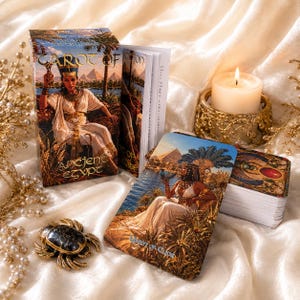Puede incluir: Una baraja de Tarot of Ancient Egypt con una caja que muestra una figura egipcia, un libro guía y una carta de la Reina de Copas. Una figura de escarabajo negro y una vela blanca en un soporte dorado se encuentran sobre una tela blanca.