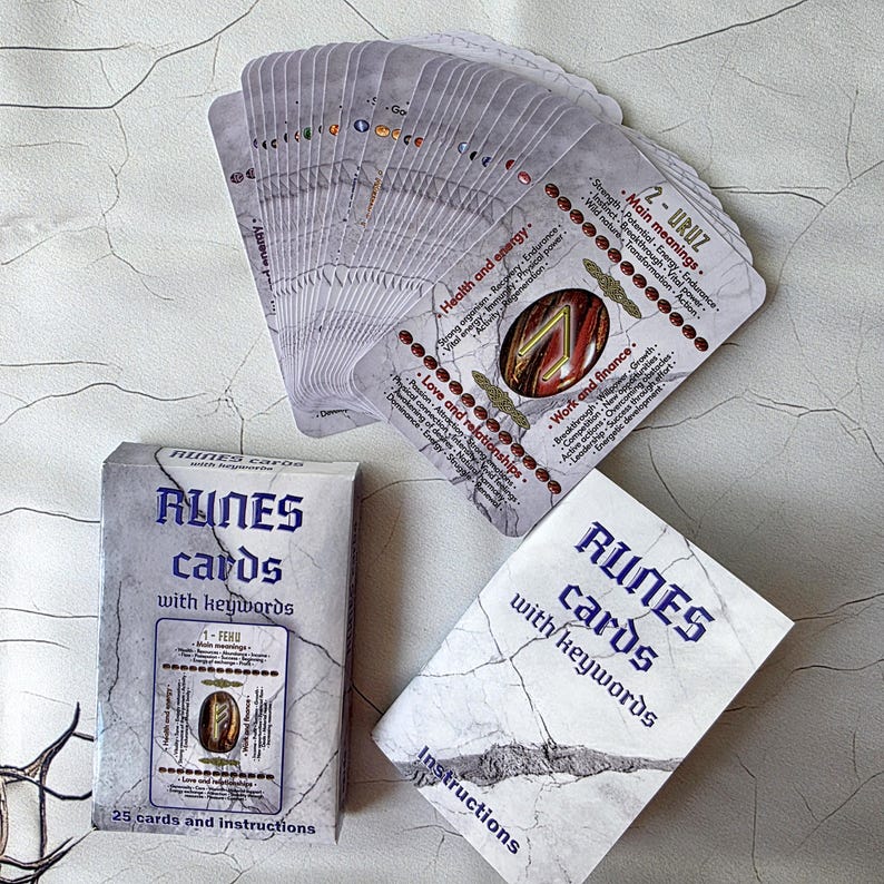 May include: A deck of rune cards with keywords, fanned out to display the card backs. The cards are white with black text and illustrations. A box and instruction card are also present, featuring the same design.