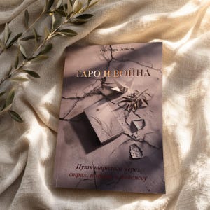 Op de afbeelding: Een boek getiteld "ТАРО И ВОЙНА" door Таромира Эстель met een omslag met een gebarsten oppervlak, een klein doosje, een origami kraanvogel en olijftakken. De ondertitel "Путь таролога через страх, потери и надежду" staat in donkerbruine letters.