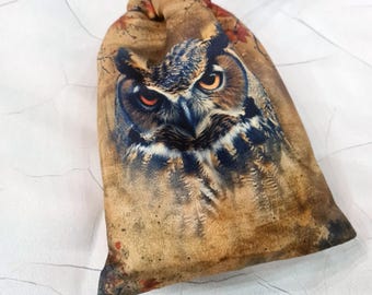 May include: A brown drawstring bag with a printed image of an owl's face. The owl has large, yellow eyes and brown feathers. The bag has a black drawstring.