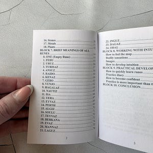 May include: Open book with a list of runes and their meanings. The pages are white with black text. The book is open to pages 2 and 3, with a hand holding the book.