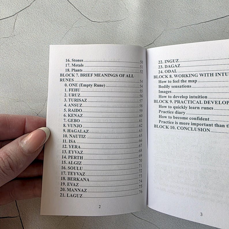 May include: Open book with a list of runes and their meanings. The pages are white with black text. The book is open to pages 2 and 3, with a hand holding the book.