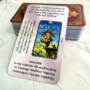 May include: A tarot card with the Fool illustration, featuring a colorful jester on a cliff edge, with text describing the card's meaning. The card is part of a deck with a decorative box.