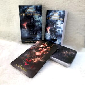 Puede incluir: Una baraja de cartas Oracle Lenormand of the Dark Archive. La caja y las cartas presentan una escena oscura y tormentosa del mar con un barco. Una carta muestra un ramo de flores. La baraja incluye 36 cartas y una guía.