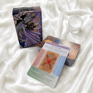 Puede incluir: Una baraja de cartas del Tarot de Thoth de Master Crowley en una caja morada con ilustraciones de hadas, un mazo de cartas, una carta titulada 2 OF WANDS con un diseño de estrella roja y texto, y un pequeño portavelas de cristal con cera blanca.