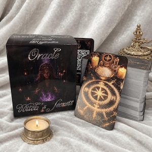 Peut inclure: Une boîte de jeu de cartes Oracle Witch's Secrets noire avec le portrait d'une personne et une lumière violette, un paquet de cartes au motif de boussole dorée, une bougie allumée dans un support doré et un objet décoratif en métal doré.