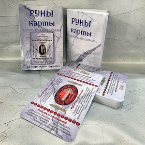 May include: A set of rune cards with accompanying booklets. The cards feature a red gemstone design with a gold arrow symbol and text in a foreign language. The booklets have a marble-like background with text in a foreign language.