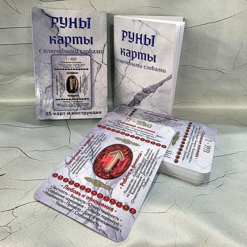 May include: A set of rune cards with accompanying booklets. The cards feature a red gemstone design with a gold arrow symbol and text in a foreign language. The booklets have a marble-like background with text in a foreign language.
