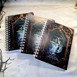May include: Three spiral-bound notebooks with ornate covers. The covers feature a dark, antique-style design with a central oval image of a tree and text in a foreign language. The notebooks are arranged on a textured surface.