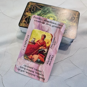 Op de afbeelding: Een tarotkaart uit een kaartspel, met een vrouw in een rode jurk die een staf vasthoudt. De kaart is gelabeld "3 - KEIZERIN" en bevat tekst over vruchtbaarheid, overvloed en liefde. Het kaartspel heeft een donkergroen en goudkleurig ontwerp.