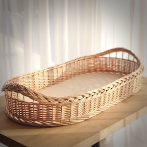 May include: A natural woven wicker basket with a rounded oval shape. The basket is made of light brown wicker and has a smooth, even weave.