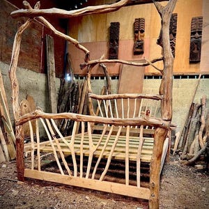 May include: A rustic, handcrafted bed frame made from natural tree branches. The bed features a canopy design and a slatted base. The wood is light brown, with visible knots and textures. The bed is in a workshop setting.