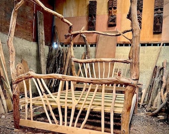 May include: A rustic, handcrafted bed frame made from natural tree branches. The bed features a canopy design and a slatted base. The wood is light brown, with visible knots and textures. The bed is in a workshop setting.