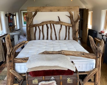 May include: A rustic wooden bed frame with a unique tree branch design. The bed is made of natural wood and features a white mattress, pillows, and blankets. Two vintage brown trunks are at the foot of the bed, with a small pink pillow on top.