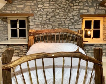 May include: A rustic wooden bed frame with a headboard and footboard made from natural branches. The bed is covered with a white comforter and pillows. The room has stone walls and small windows.