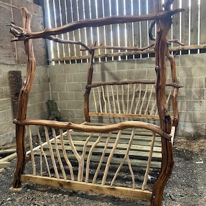 Op de afbeelding: Een rustiek, handgemaakt bedframe gemaakt van natuurlijke houten takken. Het bed heeft een hemelbedontwerp met een hoofdbord en voetbord gemaakt van verticale en horizontale houten elementen. Het frame heeft een warme bruine kleur.