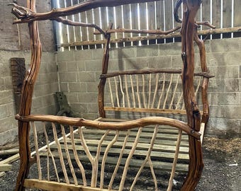 May include: A rustic, handcrafted bed frame made from natural wood branches. The bed features a canopy design with a headboard and footboard constructed from vertical and horizontal wooden elements. The frame is a warm brown color, showcasing the natural grain of the wood.