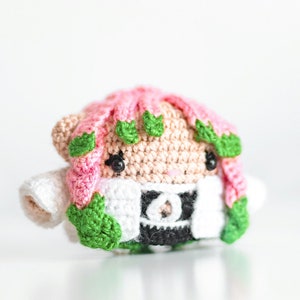 May include: A crocheted plush toy with pink hair, green leaves, and a white and black outfit. The toy has black eyes and a small, round nose.
