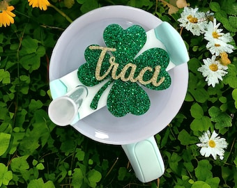 May include: A light blue tumbler with a green glitter shamrock with the name "Traci" written in gold glitter. The tumbler is surrounded by green leaves.
