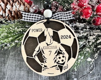May include: A wooden soccer ball ornament with a silhouette of a player in a soccer uniform. The ornament is brown with a black and white soccer ball design. The text "POWER" and "2024" are on the ornament. The player's jersey has the name "KARISSA" and the number "7" on it.