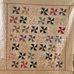 以下が含まれることがあります： ピンクの縁取りとカラフルな風車の模様が施された白いキルト。風車はさまざまな生地で作られており、グリッドパターンに配置されています。