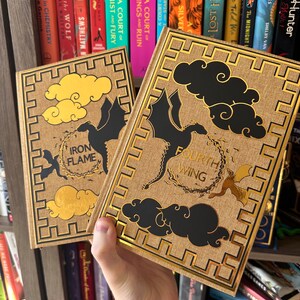 Pode incluir: Dois livros de capa dura com detalhes dourados e ilustra&ccedil;&otilde;es em preto e dourado de drag&otilde;es e nuvens. Os livros s&atilde;o intitulados "Iron Flame" e "Fourth Wing".