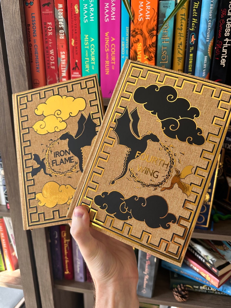 Pode incluir: Dois livros de capa dura com detalhes dourados e ilustra&ccedil;&otilde;es em preto e dourado de drag&otilde;es e nuvens. Os livros s&atilde;o intitulados "Iron Flame" e "Fourth Wing".