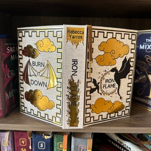 Pode incluir: Um livro branco com detalhes dourados e um design de drag&atilde;o preto e dourado. O livro &eacute; intitulado "Iron Flame" e a autora &eacute; Rebecca Yarros. O livro est&aacute; em uma estante com outros livros.