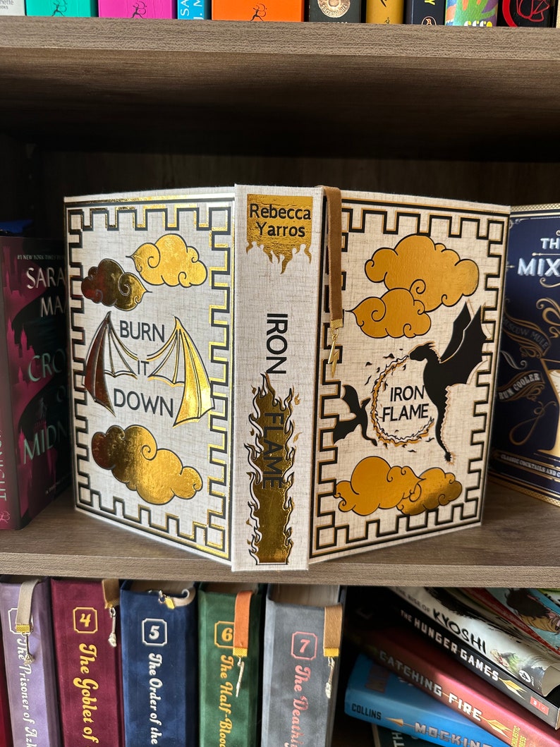 Pode incluir: Um livro branco com detalhes dourados e um design de drag&atilde;o preto e dourado. O livro &eacute; intitulado "Iron Flame" e a autora &eacute; Rebecca Yarros. O livro est&aacute; em uma estante com outros livros.