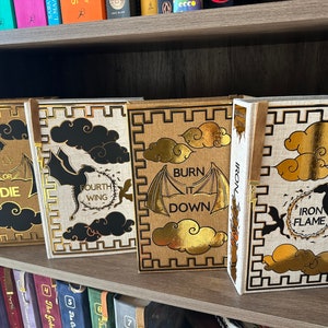 Pode incluir: Quatro livros de capa dura com desenhos dourados e pretos sobre um fundo branco. Os livros s&atilde;o intitulados "Fly or Die", "Fourth Wing", "Burn It Down" e "Iron Flame".