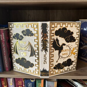 Pode incluir: Um livro de capa dura com uma capa branca e detalhes dourados. O t&iacute;tulo do livro &eacute; "Fourth Wing" e o subt&iacute;tulo &eacute; "Fly or Die". O livro est&aacute; numa estante com outros livros.