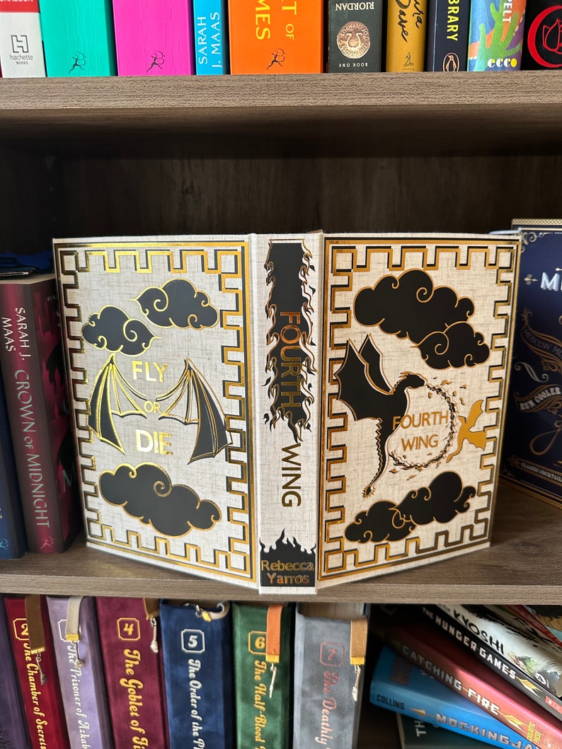 Pode incluir: Um livro de capa dura com uma capa branca e detalhes dourados. O t&iacute;tulo do livro &eacute; "Fourth Wing" e o subt&iacute;tulo &eacute; "Fly or Die". O livro est&aacute; numa estante com outros livros.