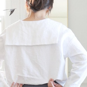 May include: A white long-sleeved top with a large, square collar. The person is wearing gray pants. The top is made of a soft fabric. The person's hair is pulled back in a bun.