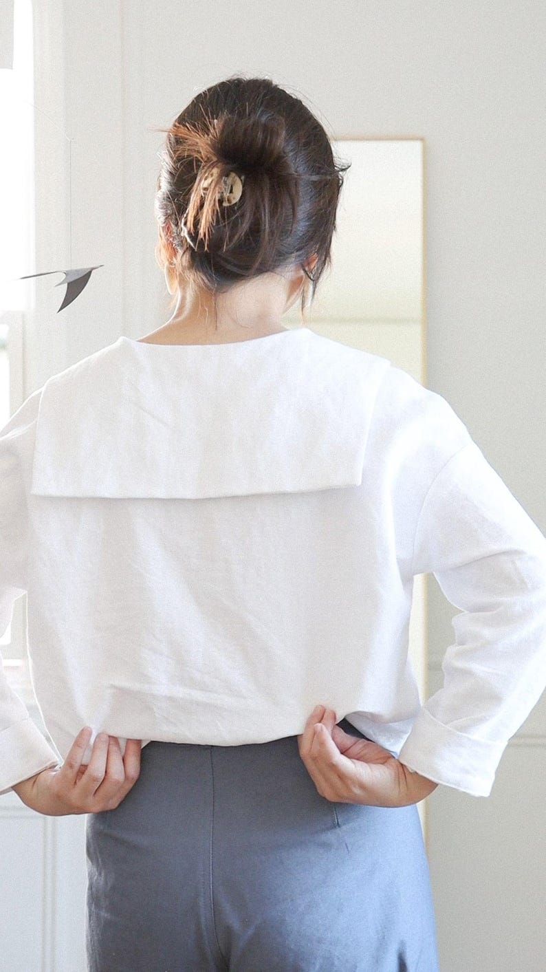 May include: A white long-sleeved top with a large, square collar. The person is wearing gray pants. The top is made of a soft fabric. The person's hair is pulled back in a bun.