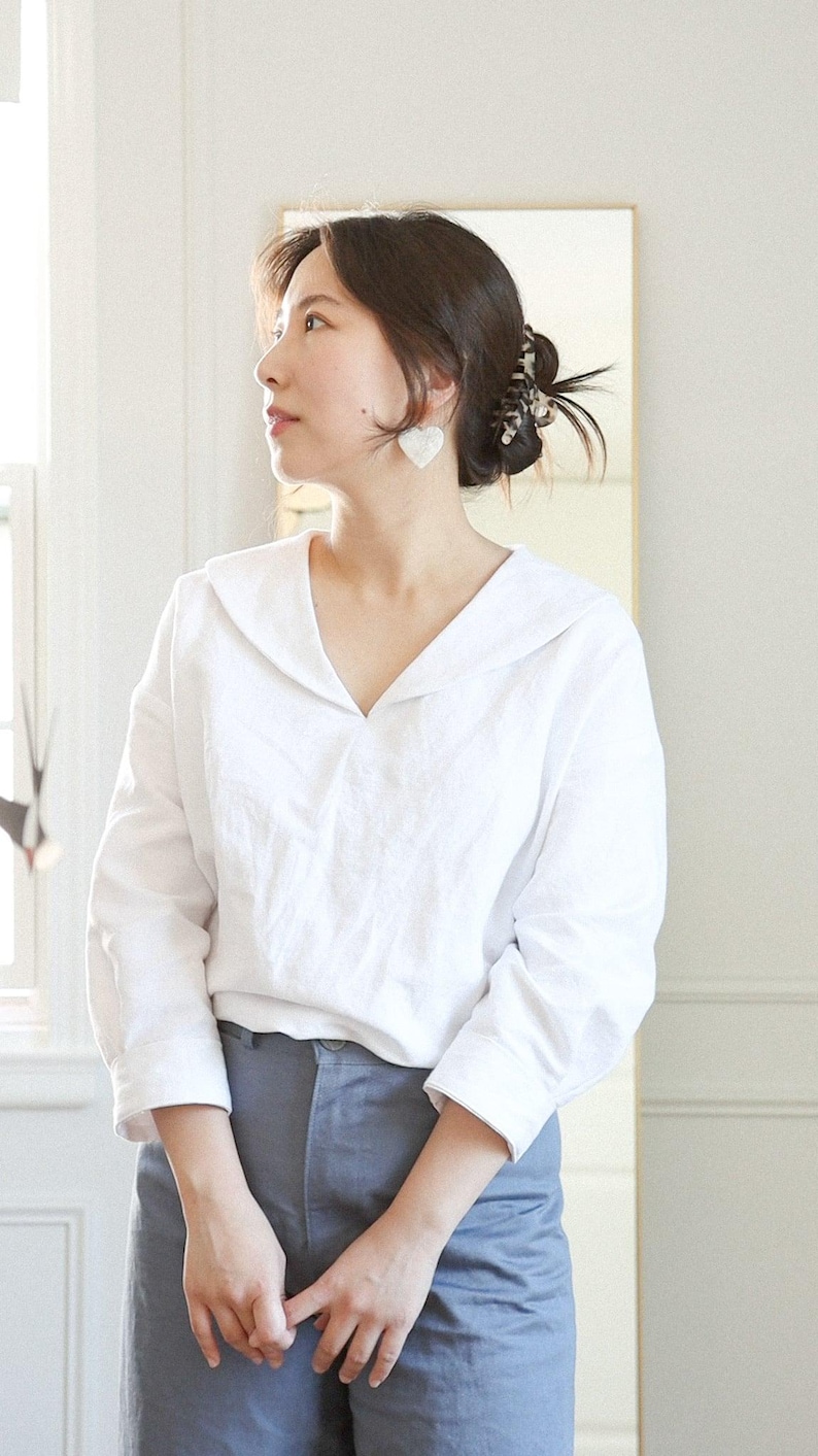 May include: A woman wearing a white linen blouse with a wide collar and long sleeves, paired with blue-grey pants. The blouse has a V-neck design. She is wearing heart-shaped earrings.