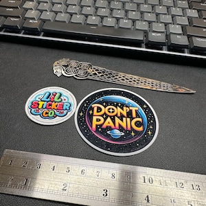 Könnte beinhalten: Zwei bunte Aufkleber auf einer schwarzen Oberfläche. Der erste Aufkleber ist rund und zeigt den Text "Lil Sticker Co." in einer Regenbogen-Schriftart. Der zweite Aufkleber ist rund und zeigt den Text "Don't Panic" in einer gelben Schriftart mit einem Planeten und Sternen im Hintergrund.