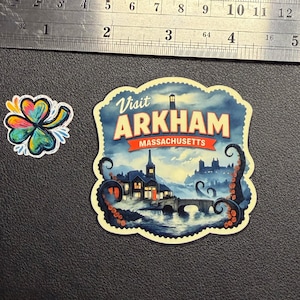 May include: Two stickers on a black surface. One is a colorful shamrock. The other is a vintage-style travel sticker that reads "Visit Arkham Massachusetts" with a spooky illustration of a town and tentacles.