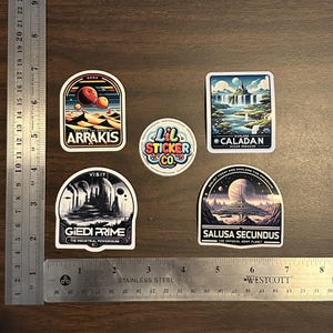 May include: Five colourful stickers with space-themed designs. The stickers feature illustrations of planets, spaceships, and landscapes. The stickers are all round or oval shaped and have white borders. The text on the stickers includes "Explore the Dunes of Arrakis", "Visit Giedi Prime", "Explore Caladan", "Salusa Secundus", and "Lil Sticker Co."