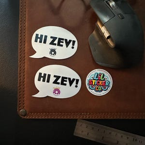 Puede incluir: Dos pegatinas blancas con forma de bocadillo con el texto "HI ZEV!" y un gráfico de donut. También es visible una pegatina redonda con el texto "Lil Sticker Co". Las pegatinas están sobre una superficie de cuero marrón.