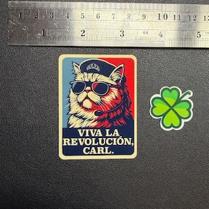 May include: A sticker with a cat wearing sunglasses, a hat, and a headset, with the text "VIVA LA REVOLUCIÓN, CARL." in a blue and red design. Beside it is a green four-leaf clover sticker. The stickers are on a dark surface.