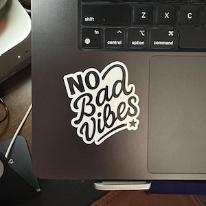 May include: A white sticker with the words "NO Bad Vibes" in a stylized font, with a small star. The sticker is on a dark gray laptop, near the keyboard.