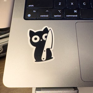 May include: A black cat sticker with a white knife graphic. The cat has large, round eyes and is holding the knife. The sticker is on a silver laptop.